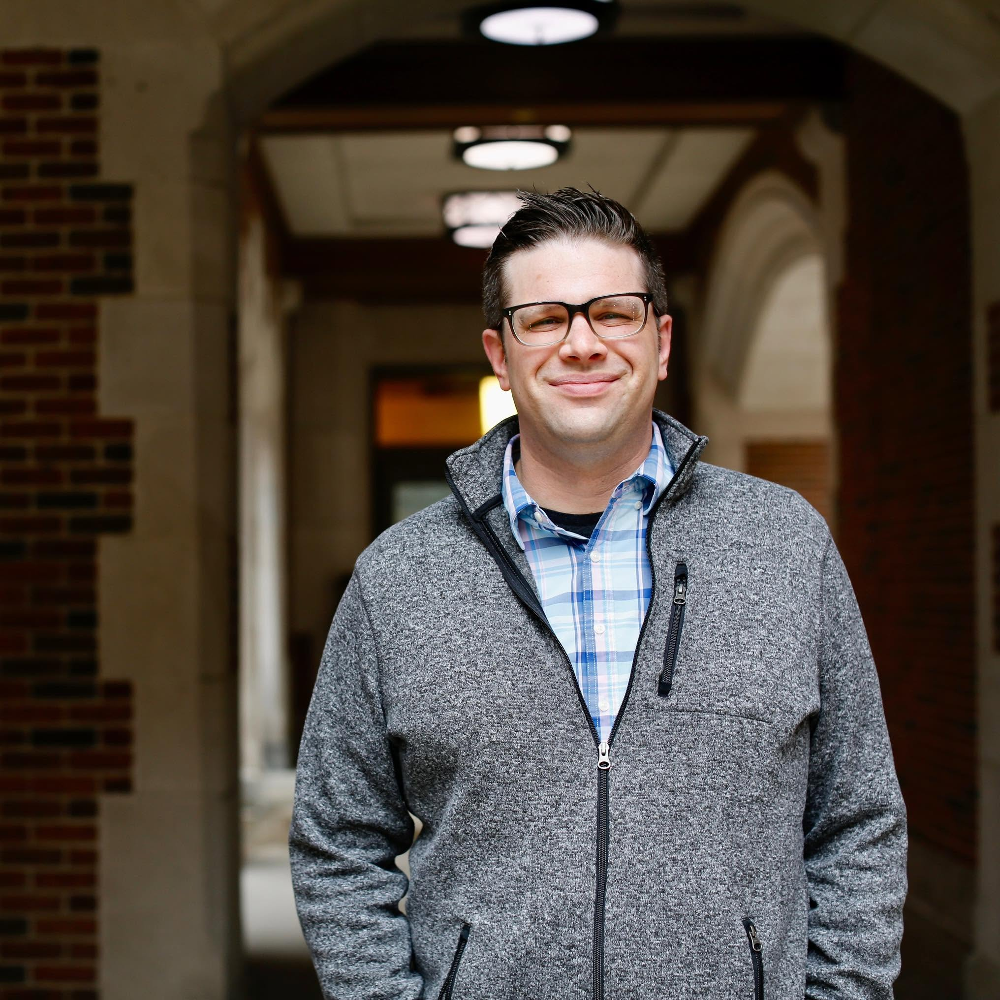 Kyle Bylin wearing glasses and a gray sweater in a brick archway